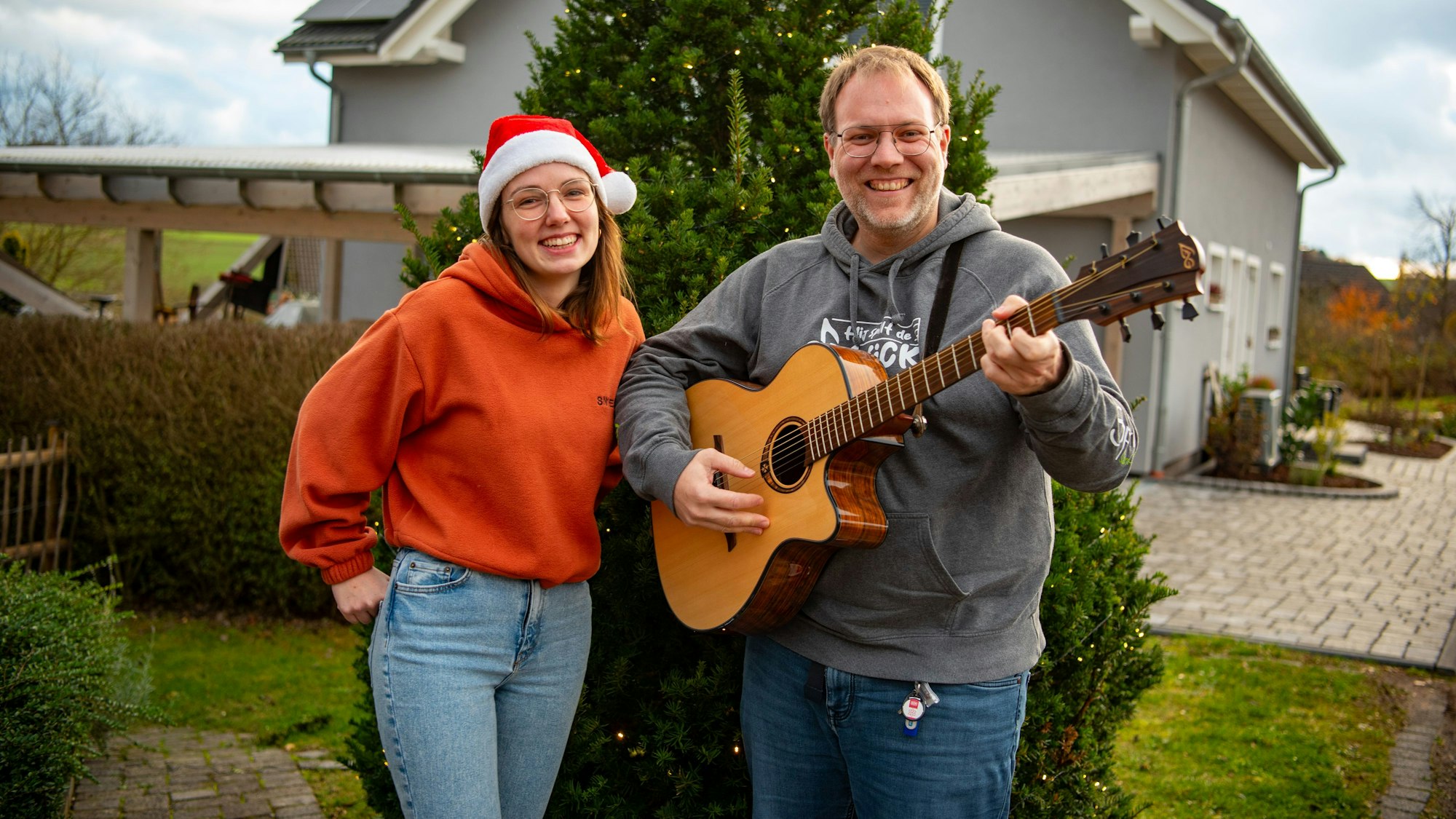 Michael Remer und Anna Jardin  von der Sacro-Pop-Band Spirit stehen in Niederbettingen vor einem beleuchteten Tannenbaum. Anna Jardin trägt eine Weihnachtsmütze, einen orangen Pullover und helle Jeans. Michael Renner trägt grauen Pullover und dunkle Jeans und spielt Gitarre.