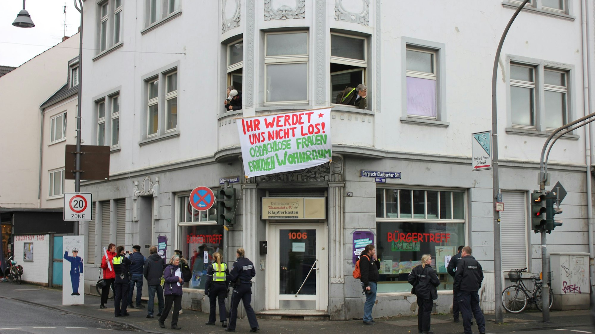 Ein Transparent mit der Aufschrift Ihr werdet uns nicht los! Obdachlose Frauen fordern Wohnraum hängt aus dem Fenster eines Eckhauses. Vor dem Haus sind einige Personen und Polizisten zu sehen.