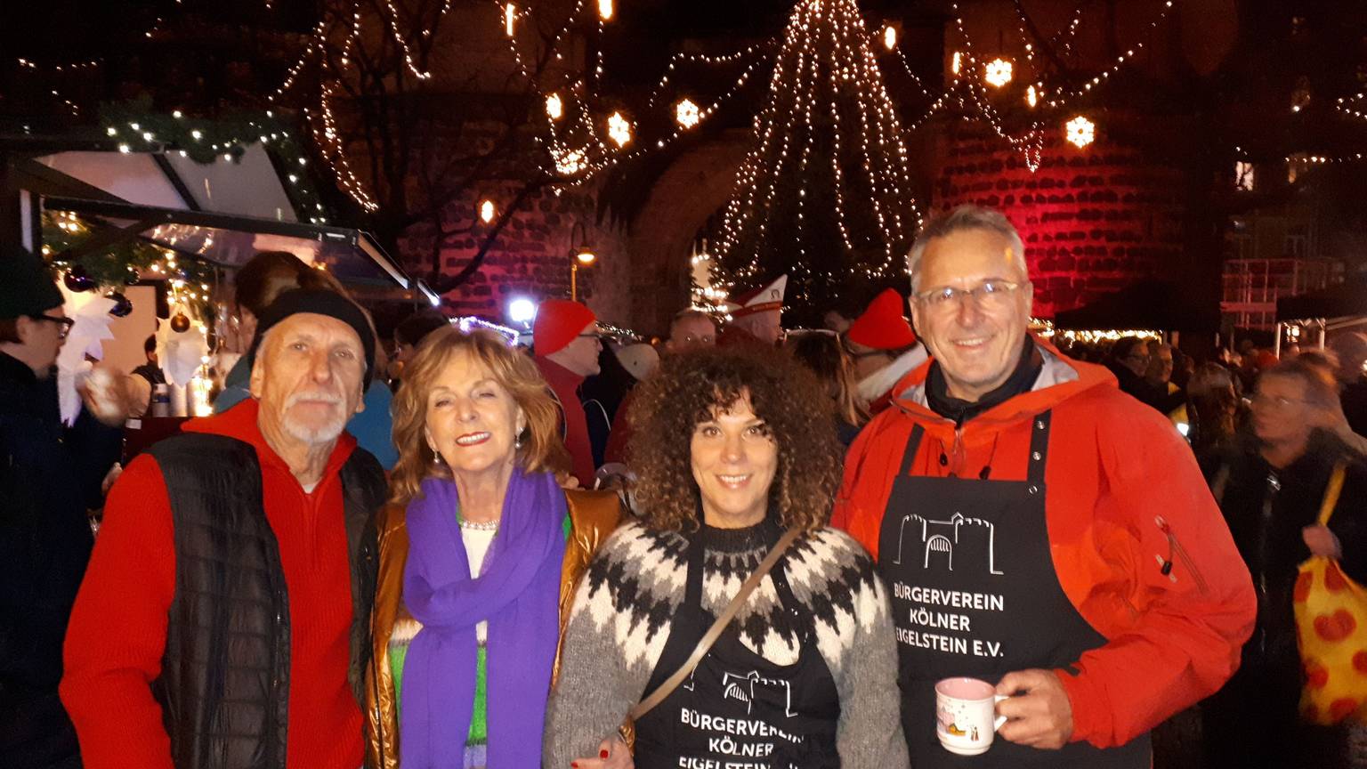 Vier Menschen stehen abends vor der festlich geschmückten Eigelsteintorburg. Im Hintergrund steht auch ein beleuchteter großer Weihnachtsbaum.