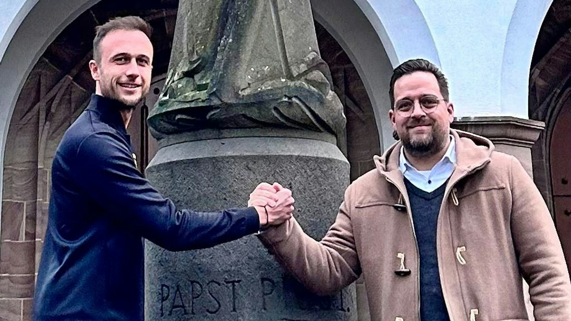 Zwei Männer stehen vor einer Statue an der Piuskirche in Zollstock und geben sich die Hände.