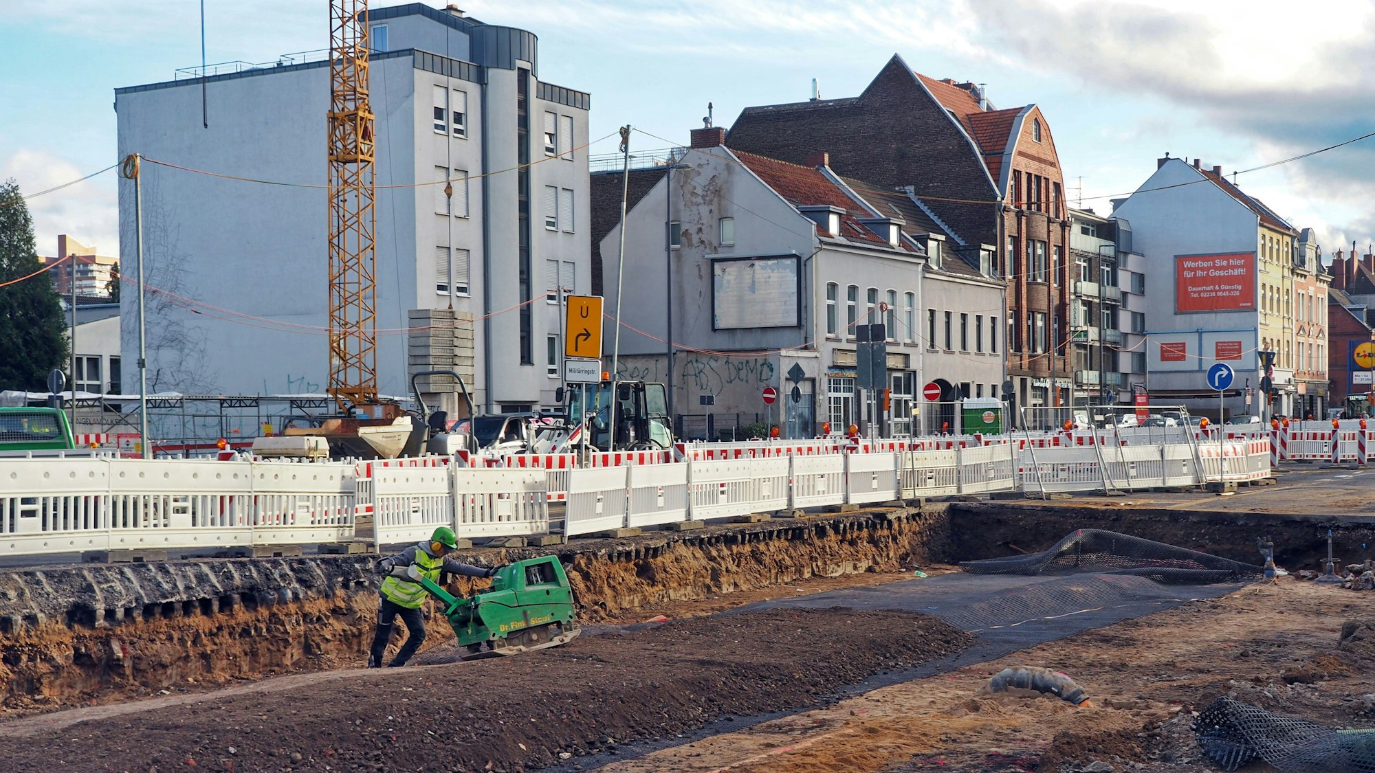 Ein Bauarbeiter arbeitet in einer Baugrube auf der Bonner Straße. Die Baugrube ist von zahlreichen rot-weißen Bauzäunen umgeben.