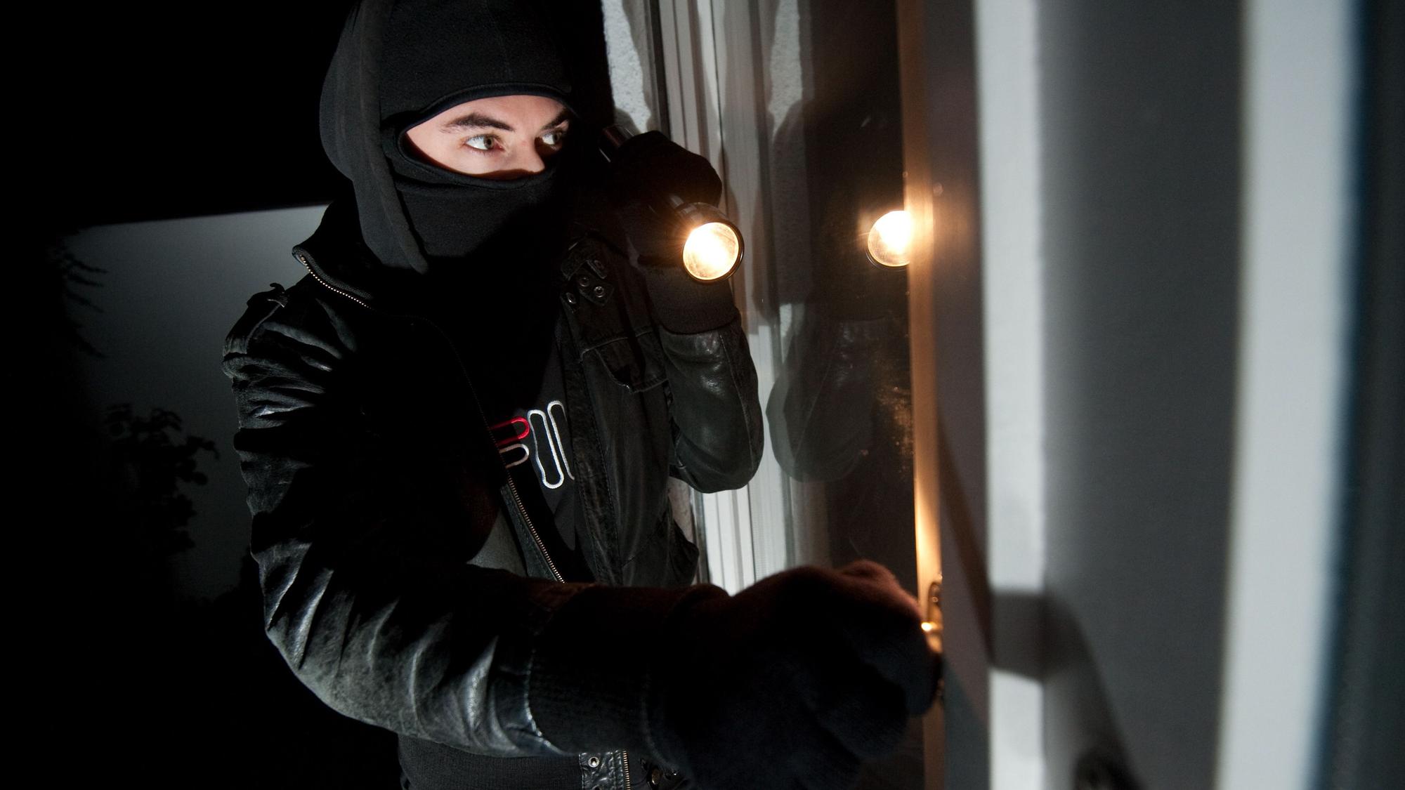 Ein vermummter Einbrecher mit Taschenlampe schaut durch ein Fenster (Symbolfoto).