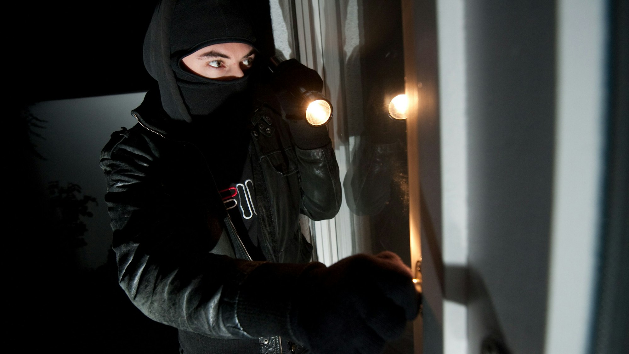 Ein vermummter Einbrecher mit Taschenlampe schaut durch ein Fenster (Symbolfoto).