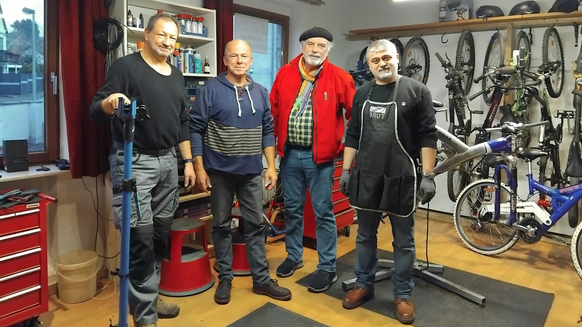 Vier Männer stehen in einer Werkstatt, an der Wand sieht man mehrere Fahrräder.