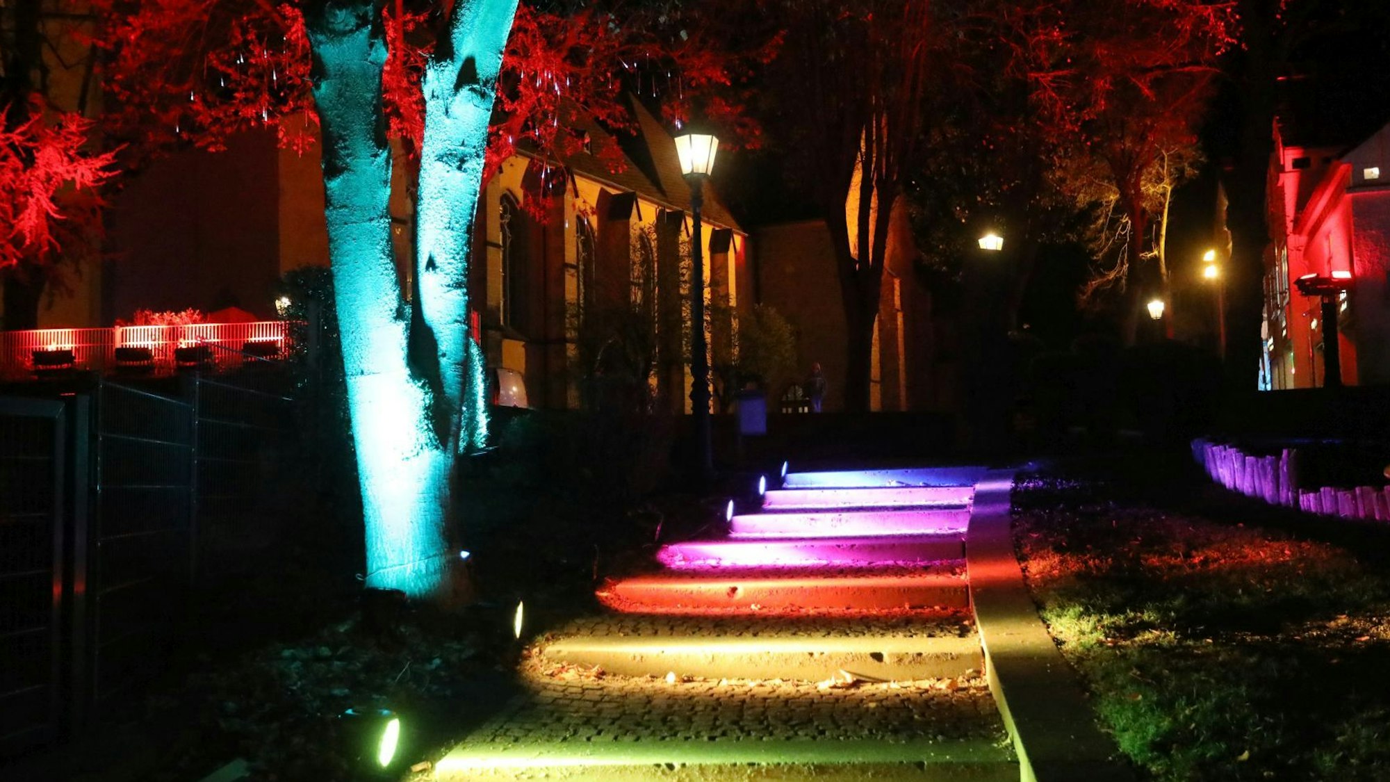 Eine Treppe, ein Baum und die Kirchenfassade sind mit bunten Lichtern angestrahlt.