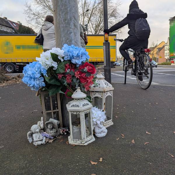 Das Foto zeigt die kleine Gedenkstätte vor dem Ampelmast an der Kreuzung, im Hintergrund ein Radfahrer und Fußgänger.