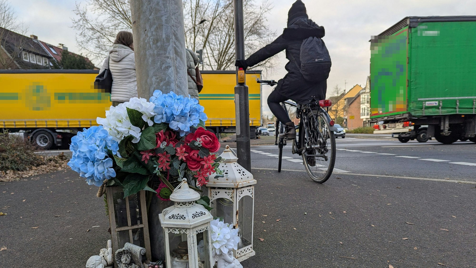 Das Foto zeigt die kleine Gedenkstätte vor dem Ampelmast an der Kreuzung, im Hintergrund ein Radfahrer und Fußgänger.