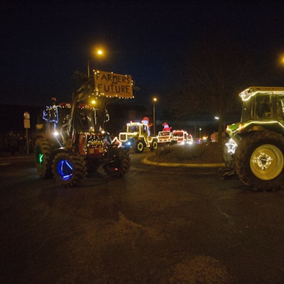 Mit bunten Lichtern geschmückte Traktoren fahren durch einen Kreisverkehr bei Nacht.