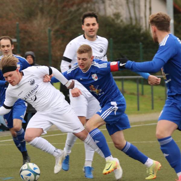 Spieler zweier Fußballmannschaften liefern sich einen Zweikampf. Der Kapitän des einen Teams drückt einen Gegner, der den Ball hat, weg.