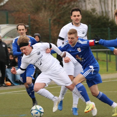 Spieler zweier Fußballmannschaften liefern sich einen Zweikampf. Der Kapitän des einen Teams drückt einen Gegner, der den Ball hat, weg.