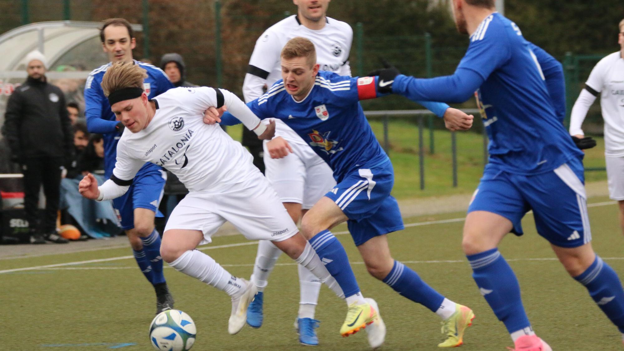 Spieler zweier Fußballmannschaften liefern sich einen Zweikampf. Der Kapitän des einen Teams drückt einen Gegner, der den Ball hat, weg.