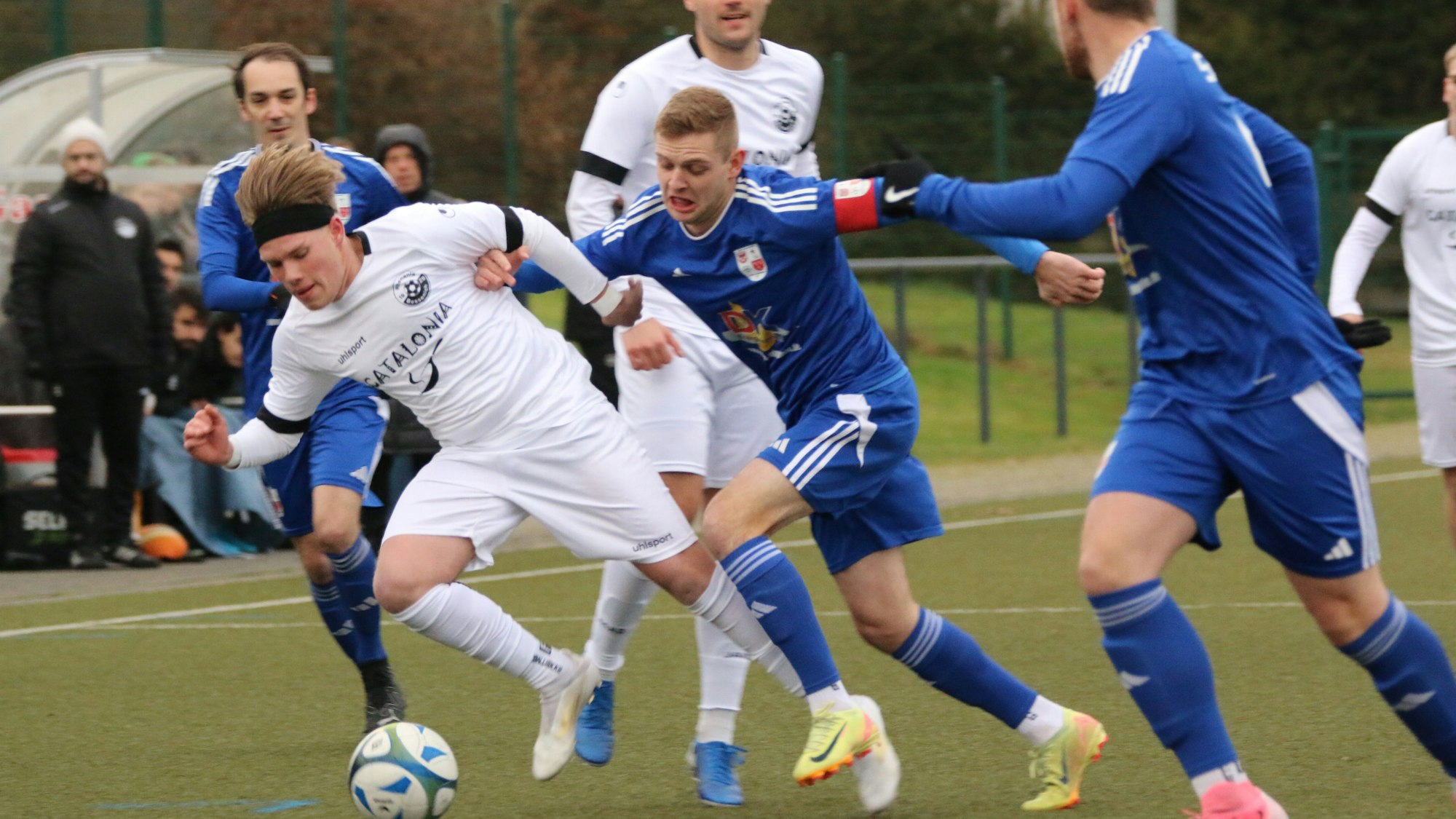 Spieler zweier Fußballmannschaften liefern sich einen Zweikampf. Der Kapitän des einen Teams drückt einen Gegner, der den Ball hat, weg.