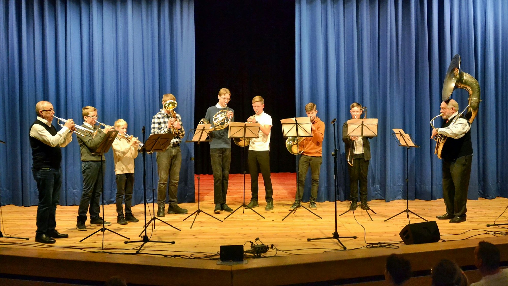 Eine Gruppe von Blasmusikern steht auf einer Bühne vor einem blauen Vorhang.