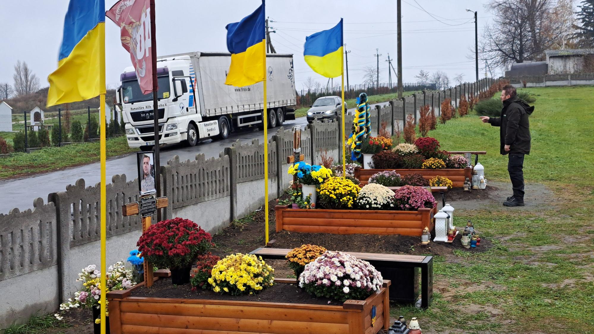 Ein Mann steht vor Gräbern mit ukrainischen Fahnen, im Hintergrund ist ein Lkw der Humanitären Hilfe Overath zu sehen.