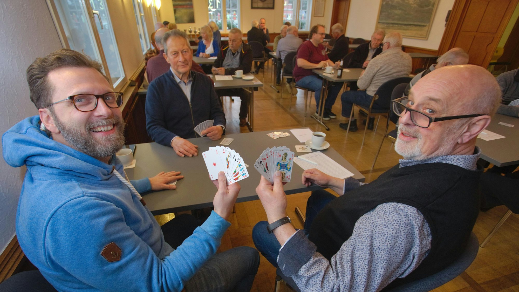 Drei Männer sitzen an einem Tisch und halten Spielkarten in Händen, im Hintergrund sind weitere Tische mit Spielern zu sehen.