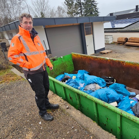 Der Bauhofleiterin orangefarbener Warnjacke steht neben einem grünen Container voller Müllsäcke.
