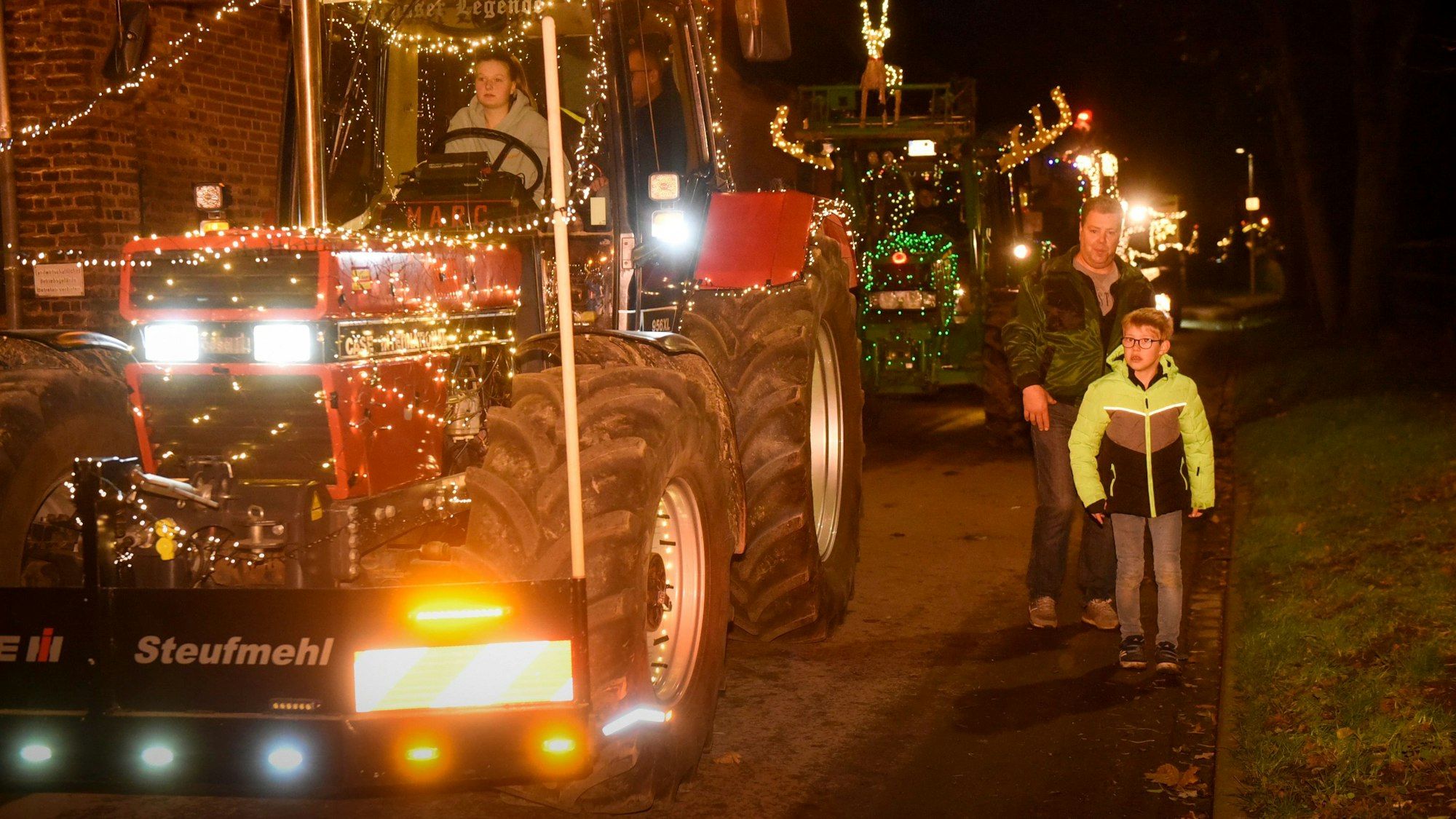 Auf dem Foto sind mit Lichterketten geschmückte Traktoren zu sehen. Menschen stehen am Wegesrand und verfolgen den Zug.