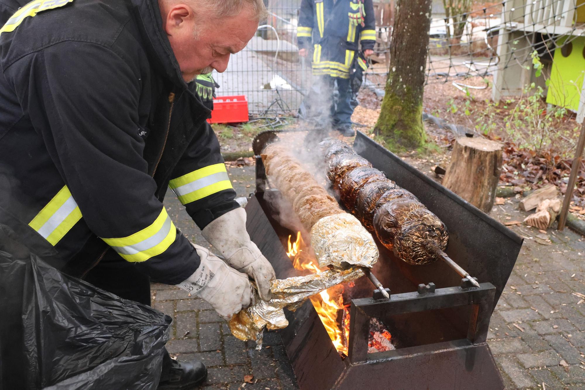 Heinz-Josef Stangier trägt eine schwarze Feuerwehrjacke und dicke Handschuhe und befreit den Spießbraten von seiner Umhüllung aus Alu-Folie. Darunter prasseln Flammen auf einem Grill.