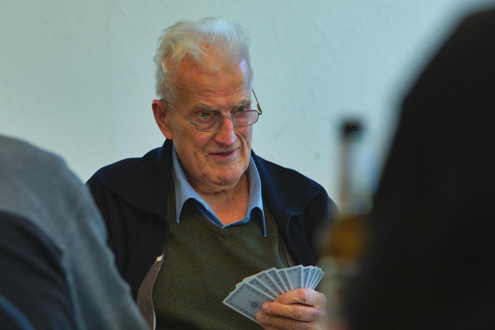 Ein Mann mit weißen Haaren sitzt an einem Tisch und hält Spielkarten in der Hand.