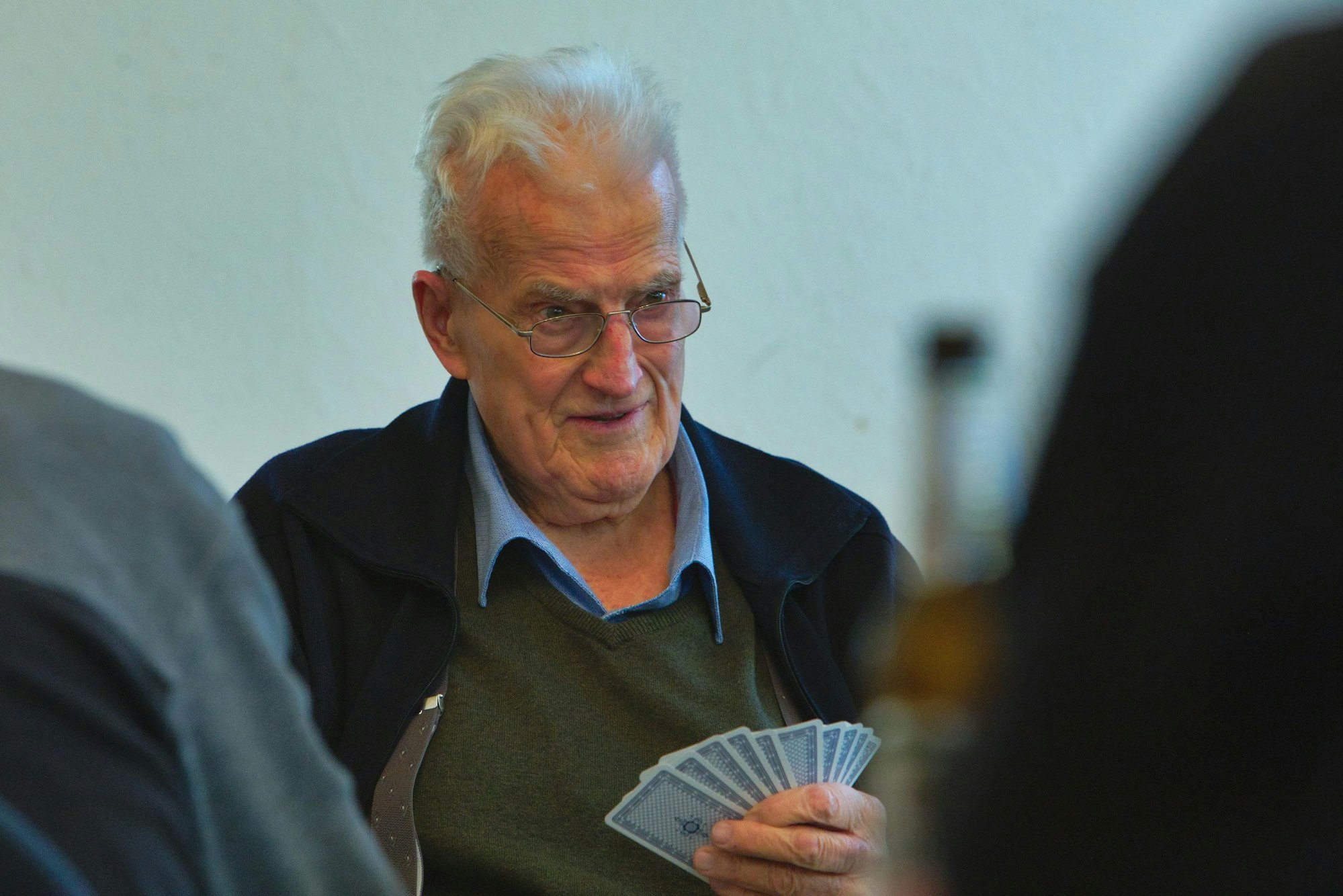 Ein Mann mit weißen Haaren sitzt an einem Tisch und hält Spielkarten in der Hand.