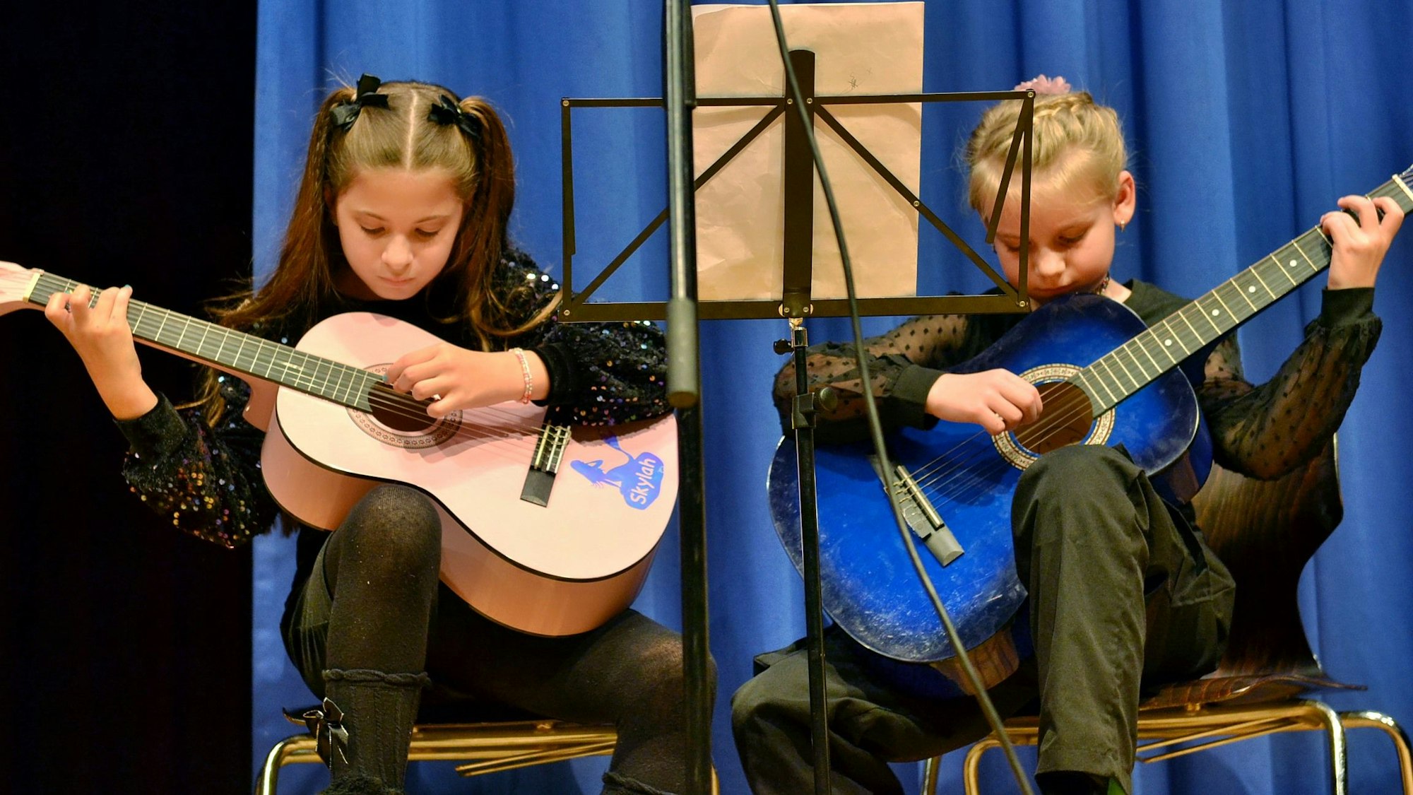Zwei Mädchen sitzen in schwarzer Kleidung vor einem Notenständer und spielen Gitarre.