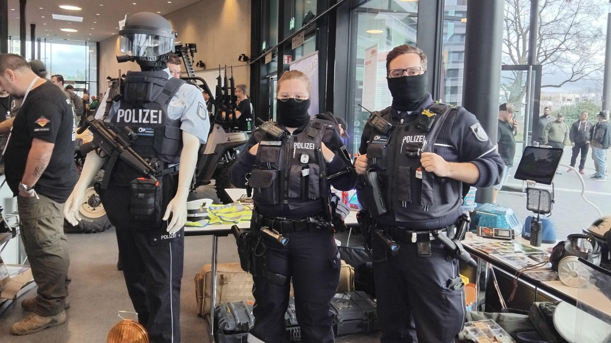 Eine Frau und ein Mann in Polizeiuniformen stehen vor einem Messestand, neben ihnen eine uniformierte Schaufensterpuppe, die Waffen und einen Helm trägt.