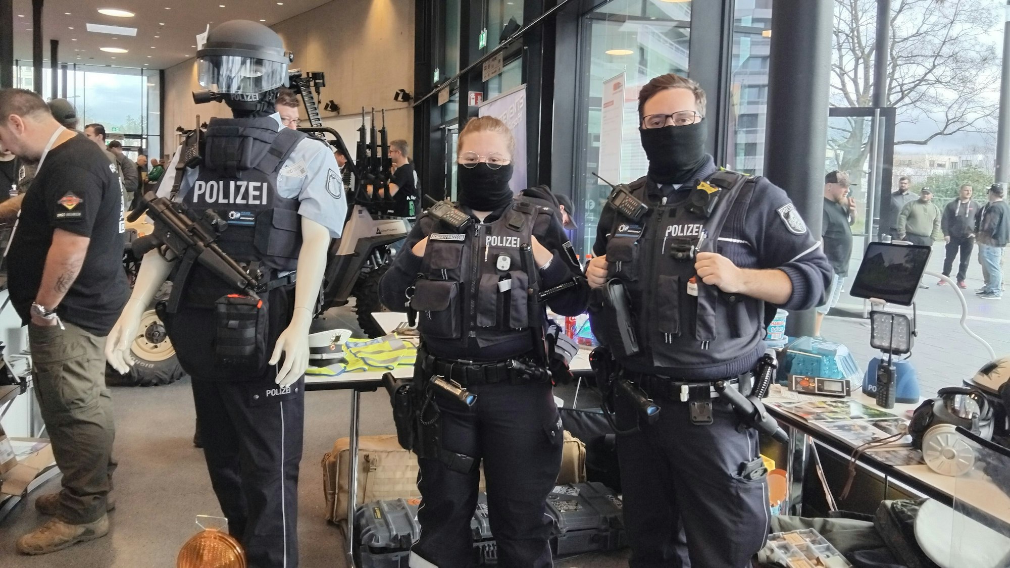 Eine Frau und ein Mann in Polizeiuniformen stehen vor einem Messestand, neben ihnen eine uniformierte Schaufensterpuppe, die Waffen und einen Helm trägt.