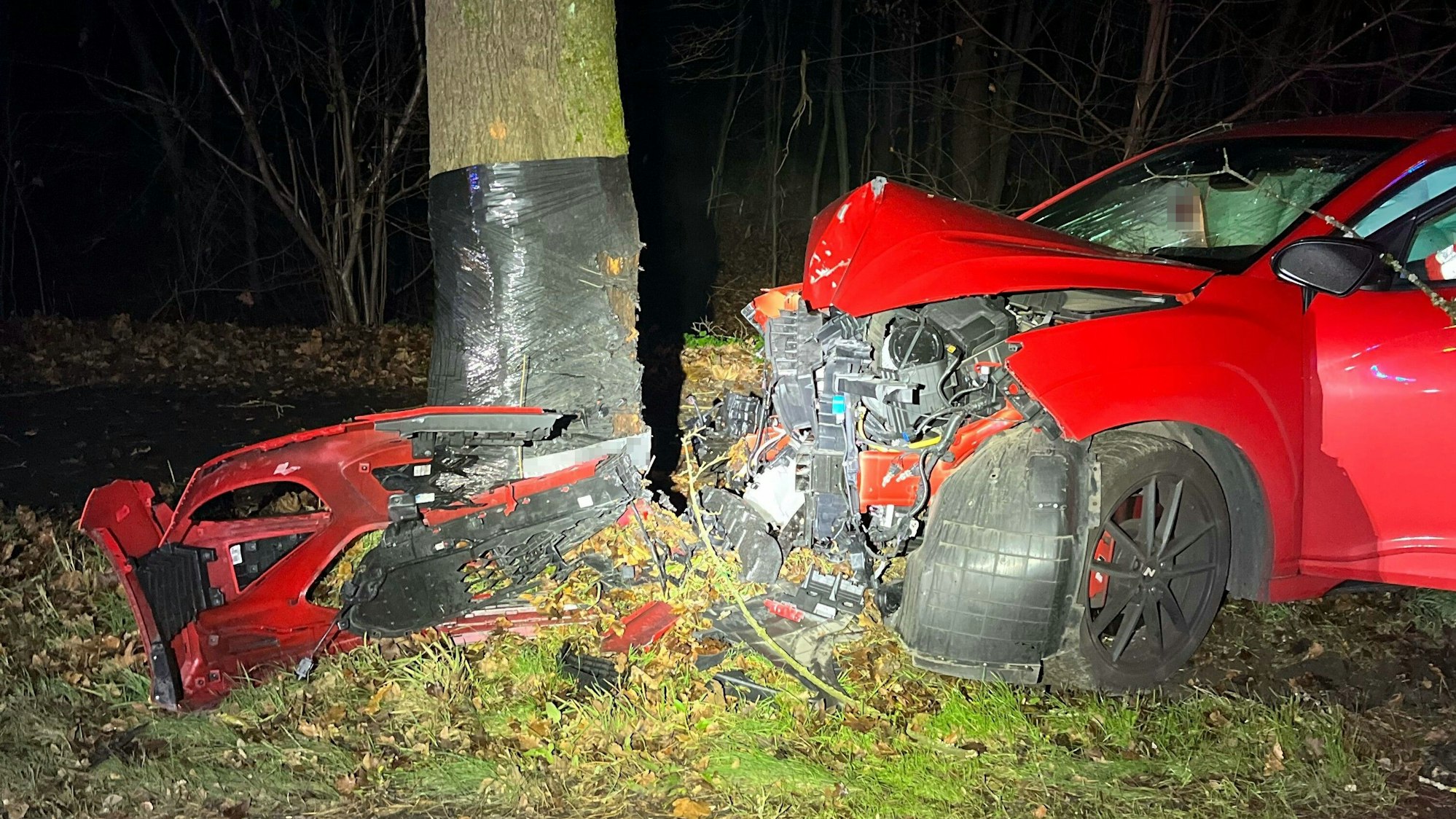 Ein rotes Auto steht schwer beschädigt vor einem Baum, gegen den es gerade gefahren ist.
