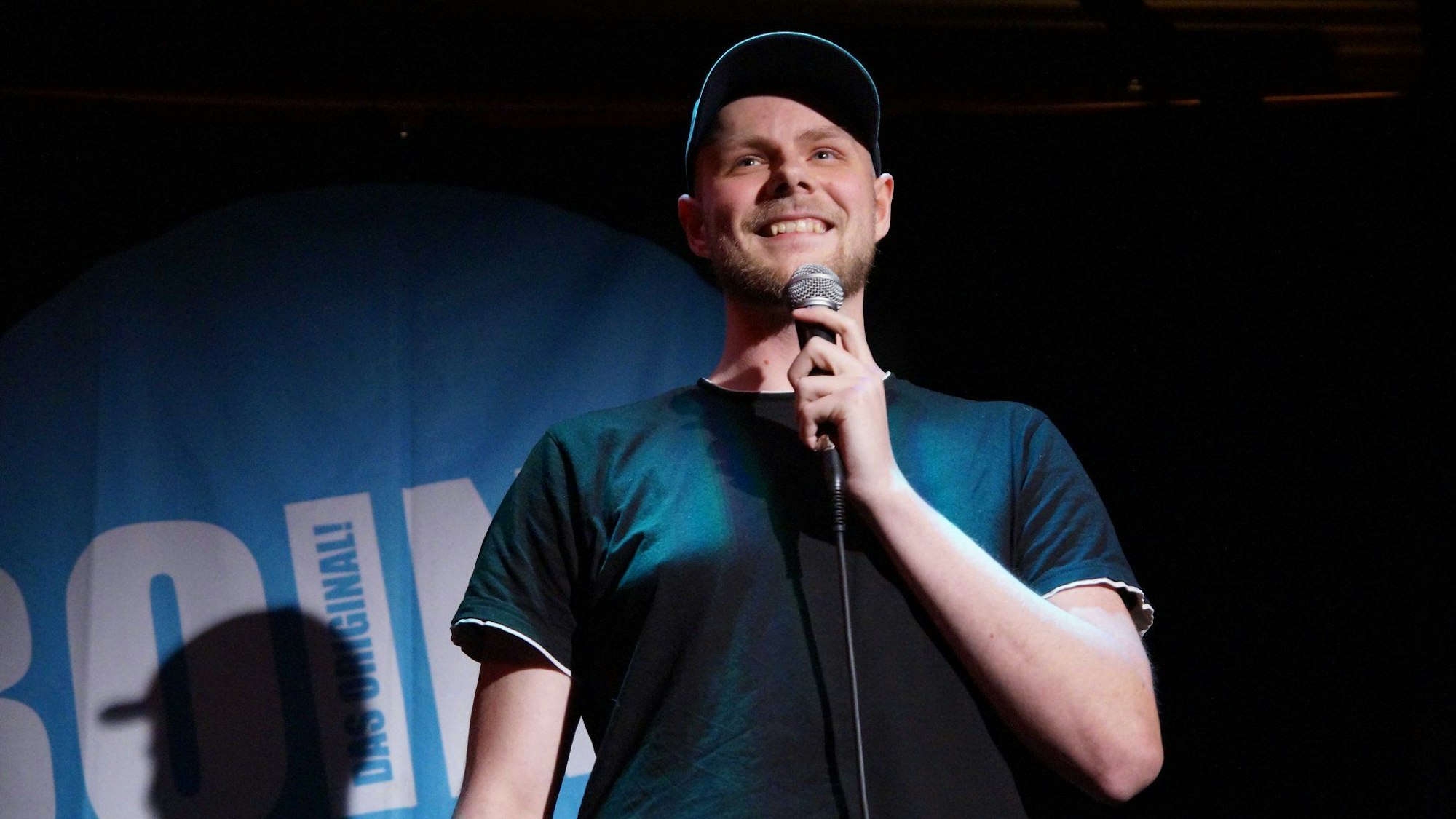 Ein Mann steht lächelnd auf einer Bühne, Scheinwerfer sind auf ihn gerichtet, er hält ein Mikrophon.