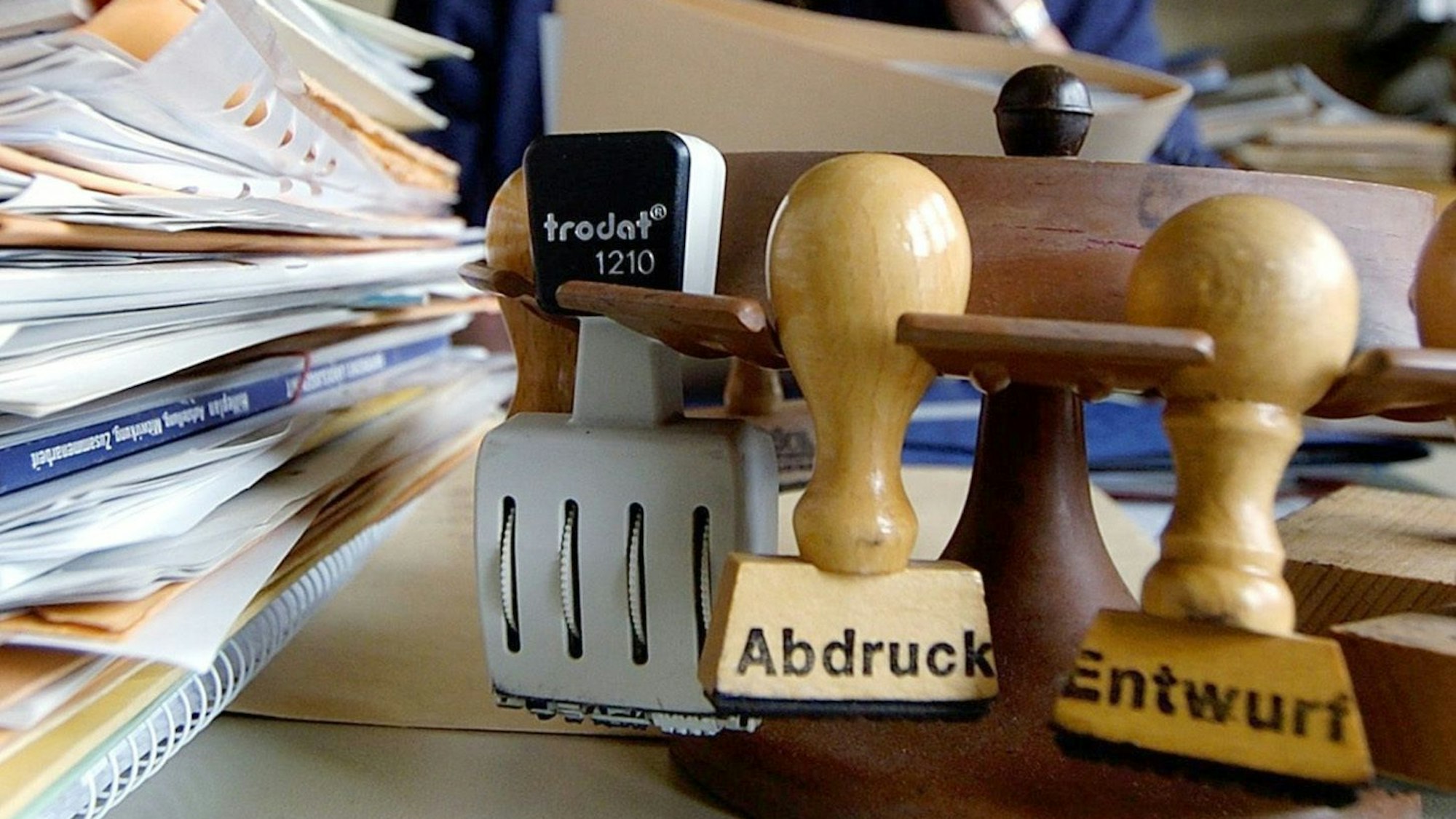 Eine Mitarbeiterin eines Landratsamts sitzt telefonierend hinter zahlreichen Akten und Stempeln an ihrem Arbeitsplatz.