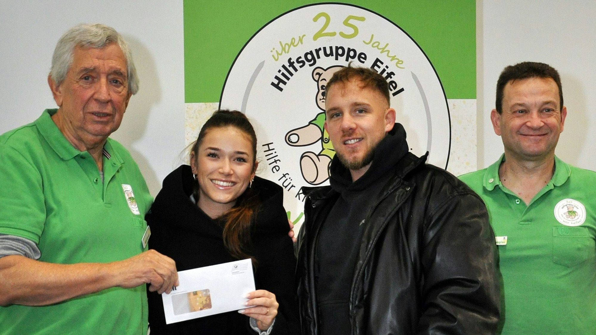 Drei Männer und eine Frau stehen nebeneinander. Die Frau überreicht ihrem Nebenmann, im Bild links, ein Kuvert.