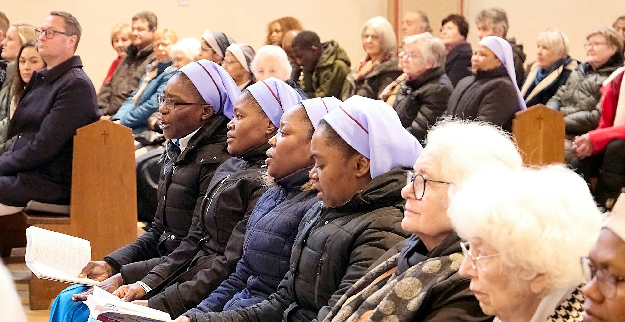 Vier afrikanische Schwestern sitzen mit ihren Gesangbüchern nebeneinander auf einer der Kirchenbänke, daneben und dahinter weitere Besucher des Gottesdienstes.