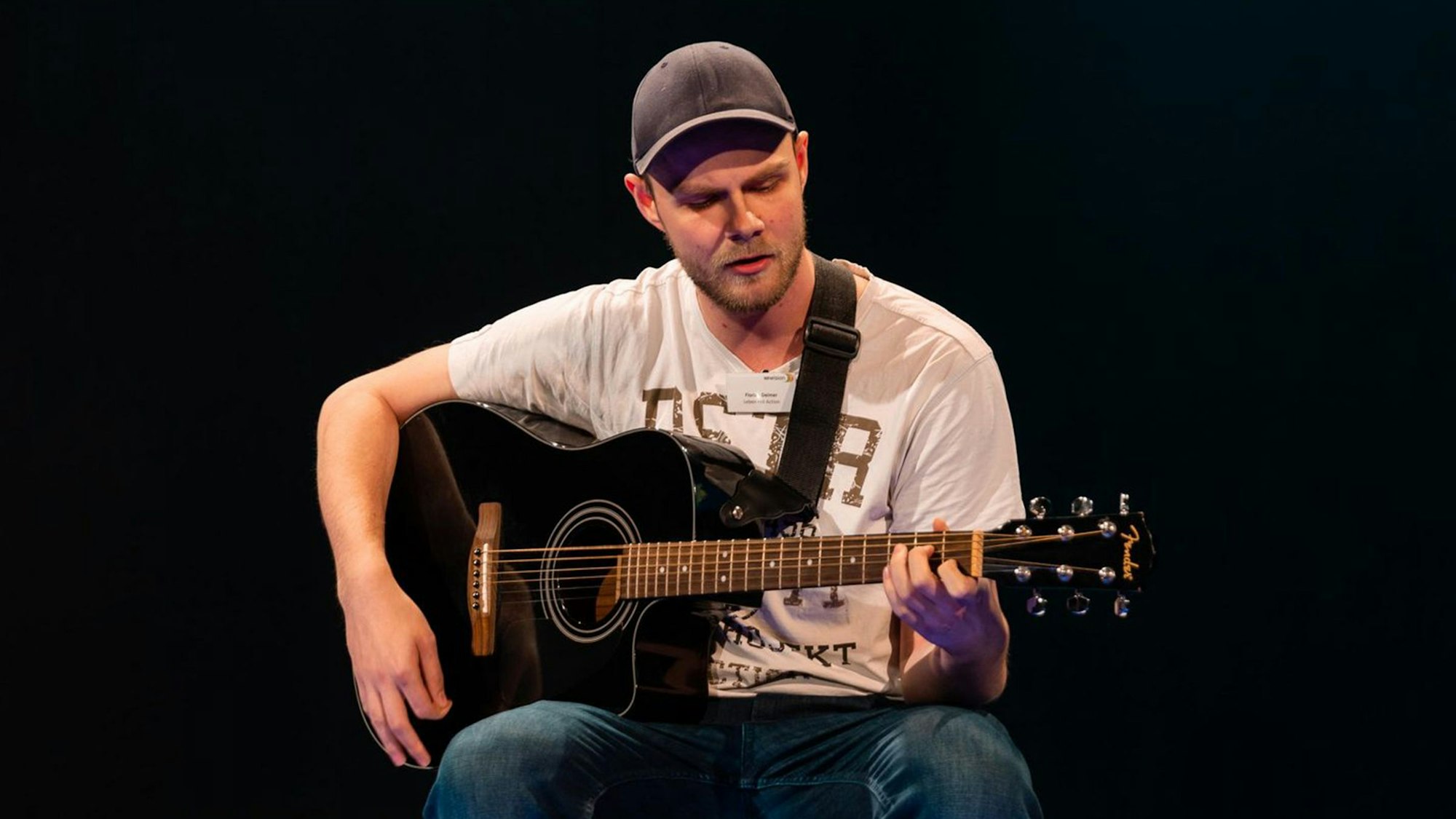 Ein Mann sitzt auf einer Bühne, der Hintergrund ist schwarz, er singt und spielt Gitarre.