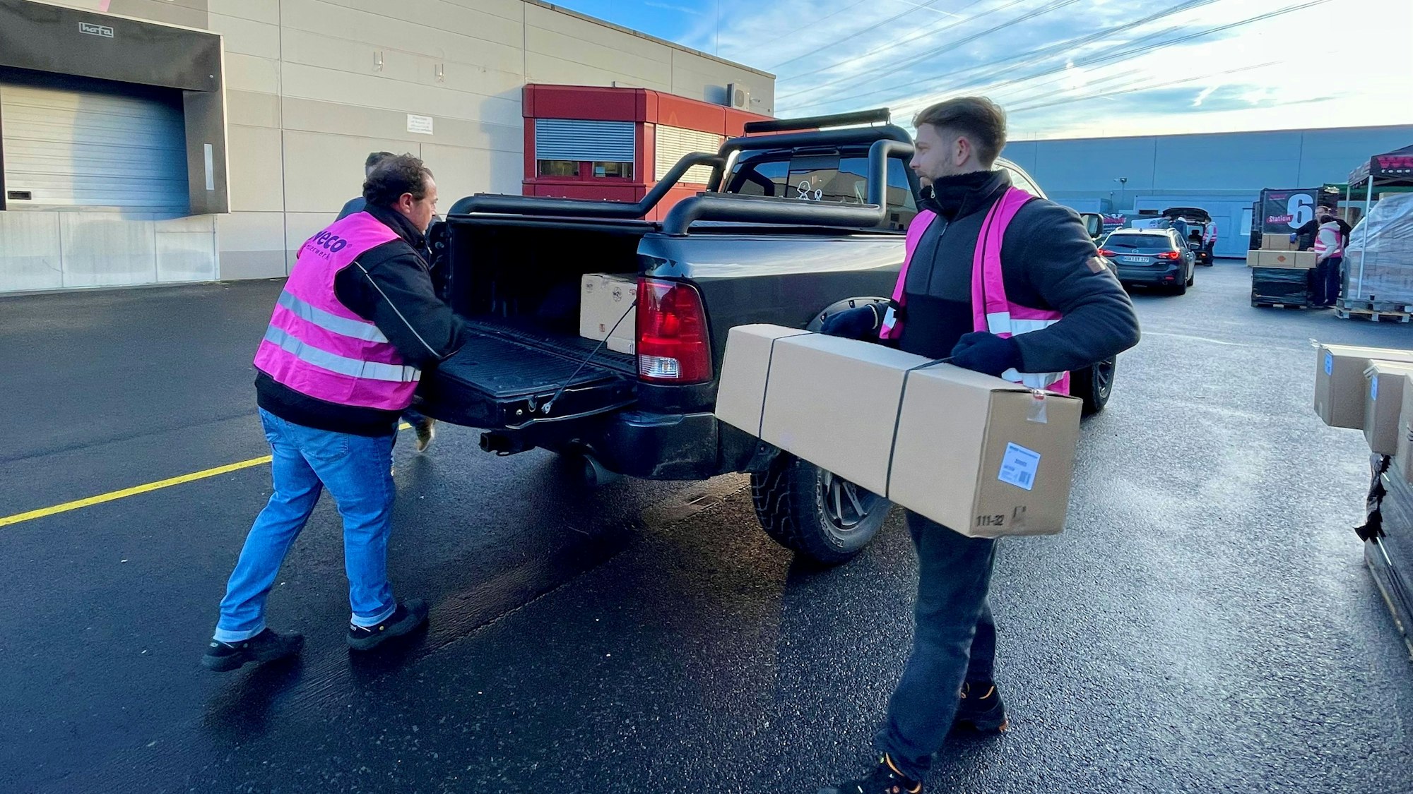 Zwei Männer in pinkfarbenen Weco-Westen heben große Pakete auf die Ladefläche eines Pick-ups.