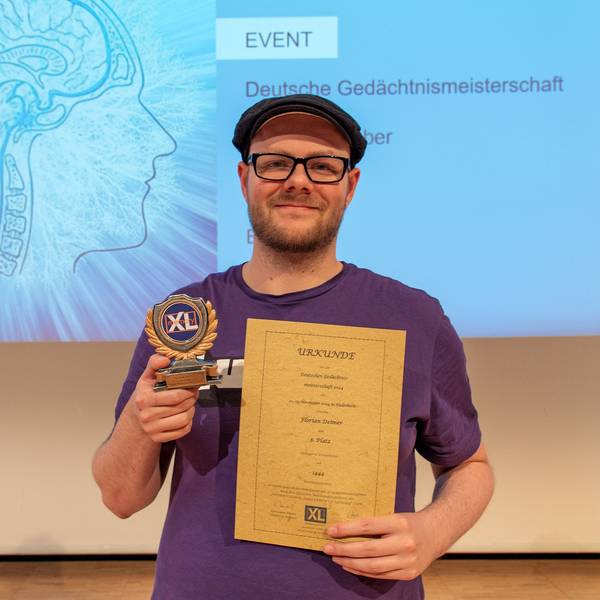 Ein Mann hält eine Trophäe und eine Urkunde in der Hand, hinter ihm ist eine Projektion mit einem Gehirn-Symbol und dem Titel „Deutsche Gedächtnismeisterschaft“ zu sehen.