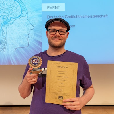 Ein Mann hält eine Trophäe und eine Urkunde in der Hand, hinter ihm ist eine Projektion mit einem Gehirn-Symbol und dem Titel „Deutsche Gedächtnismeisterschaft“ zu sehen.