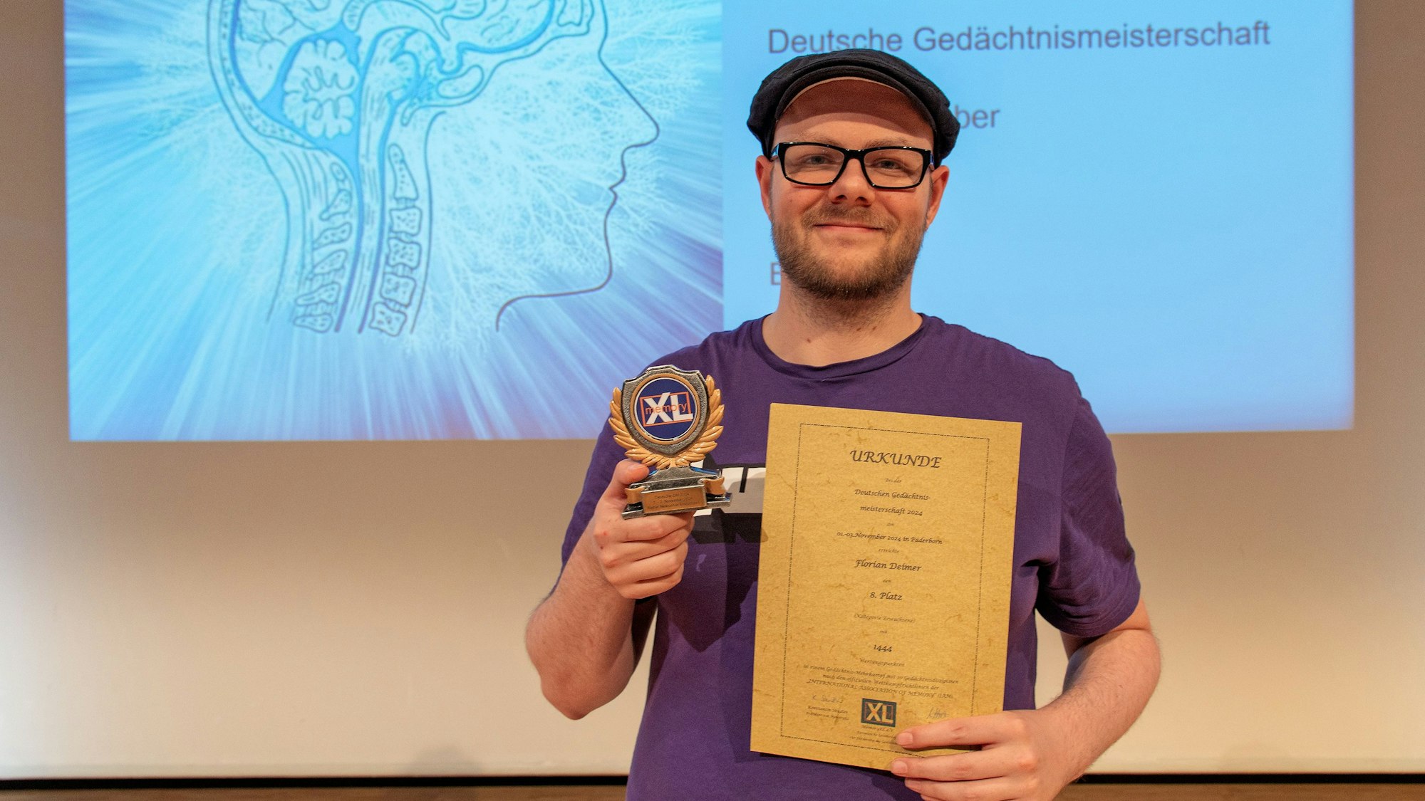 Ein Mann hält eine Trophäe und eine Urkunde in der Hand, hinter ihm ist eine Projektion mit einem Gehirn-Symbol und dem Titel „Deutsche Gedächtnismeisterschaft“ zu sehen.