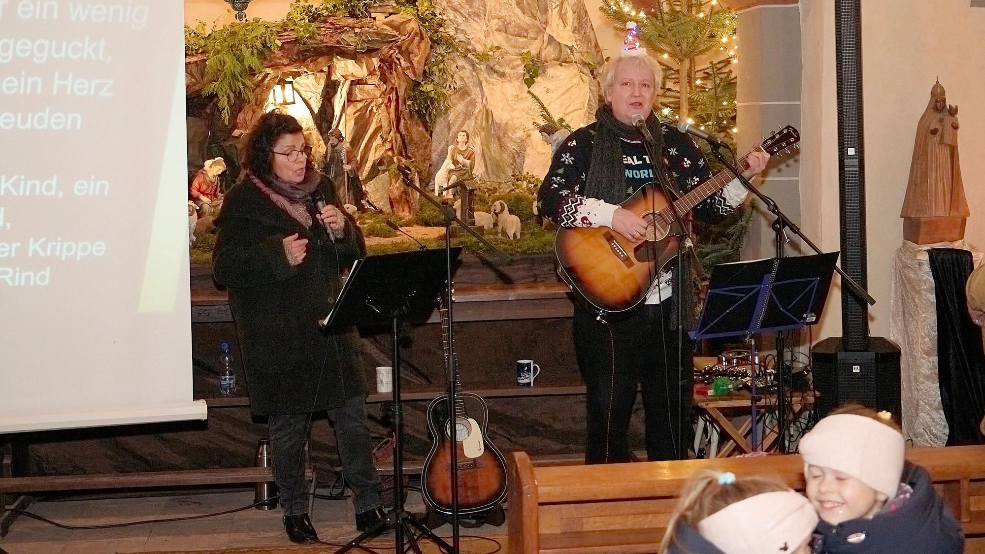 Eine Frau mit Mikrophon in der Hand und ein Mann, der Gitarre spielt, stehen im Altarraum einer Kirche. Hinter ihnen ist eine Weihnachtskrippe aufgebaut.