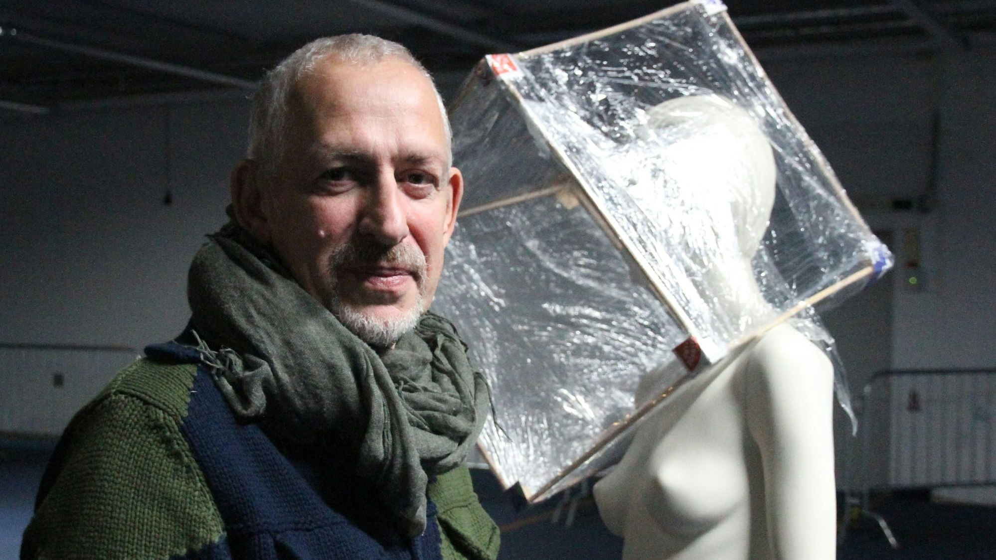 Ein Mann neben einer Schaufensterfigur, deren Kopf in einem Zellophanwürfel steckt.