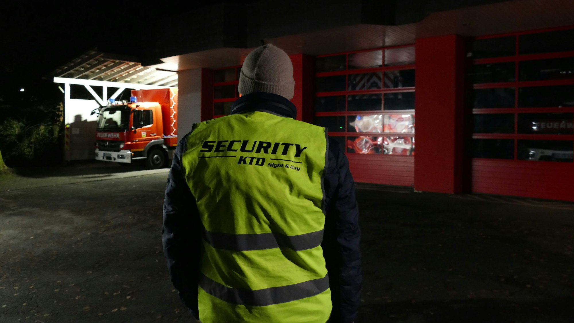 Ein Mann mit einer neongelben Weste, die mit dem Wort Security beschriftet ist, steht mit dem Rücken zur Kamera vor einem Feuerwehrgerätehaus.