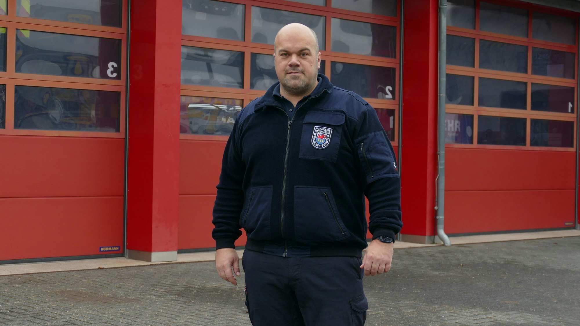 Ein Mann in einer Feuerwehrjacke blickt in die Kamera, hinter ihm sind die geschlossenen Garagentore einer Feuerwache zu sehen.