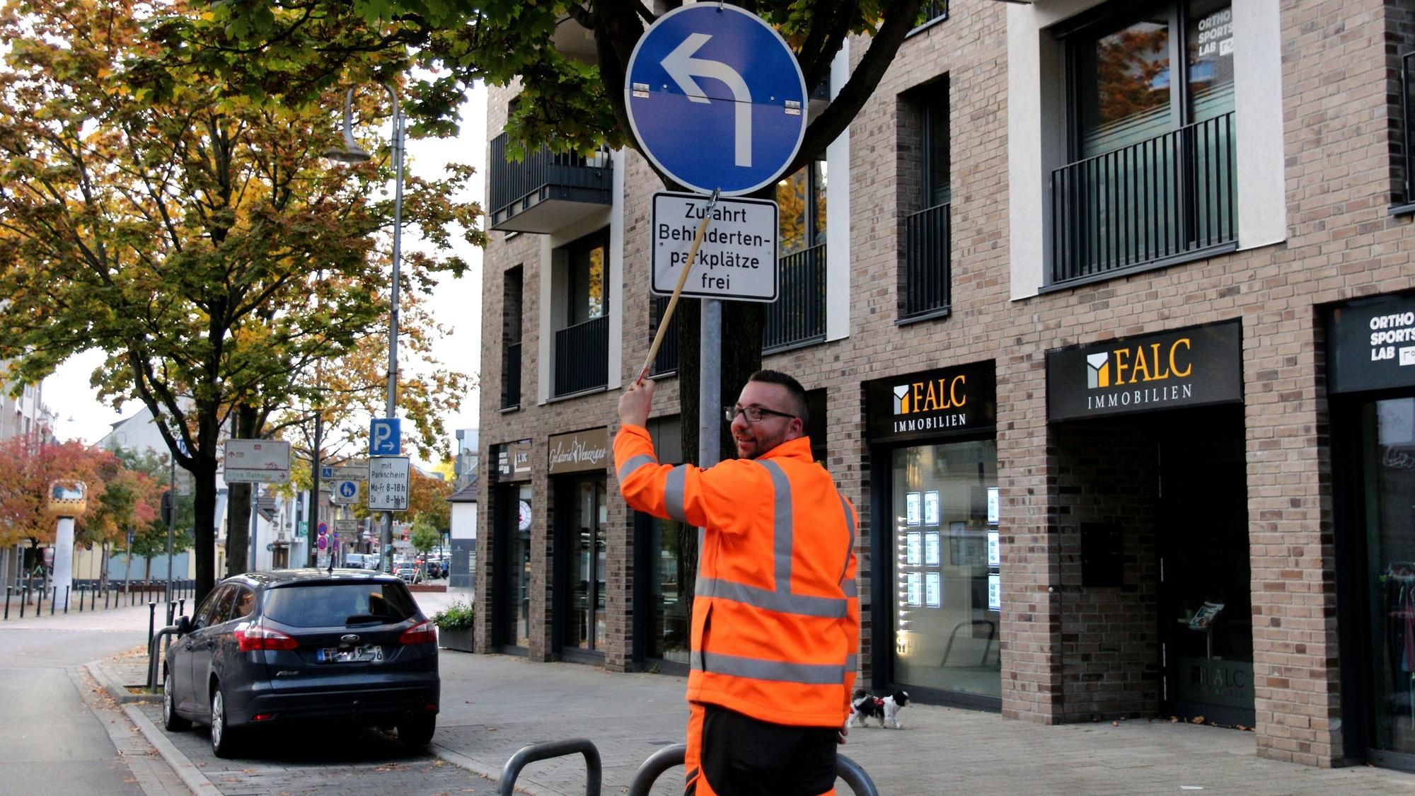 Ein Mann in orangefarbener Sicherheitsjacke klappt ein Verkehrsschild herunter. Zu lesen ist der Hinweis „Zufahrt Behindertenparkplätze frei“.