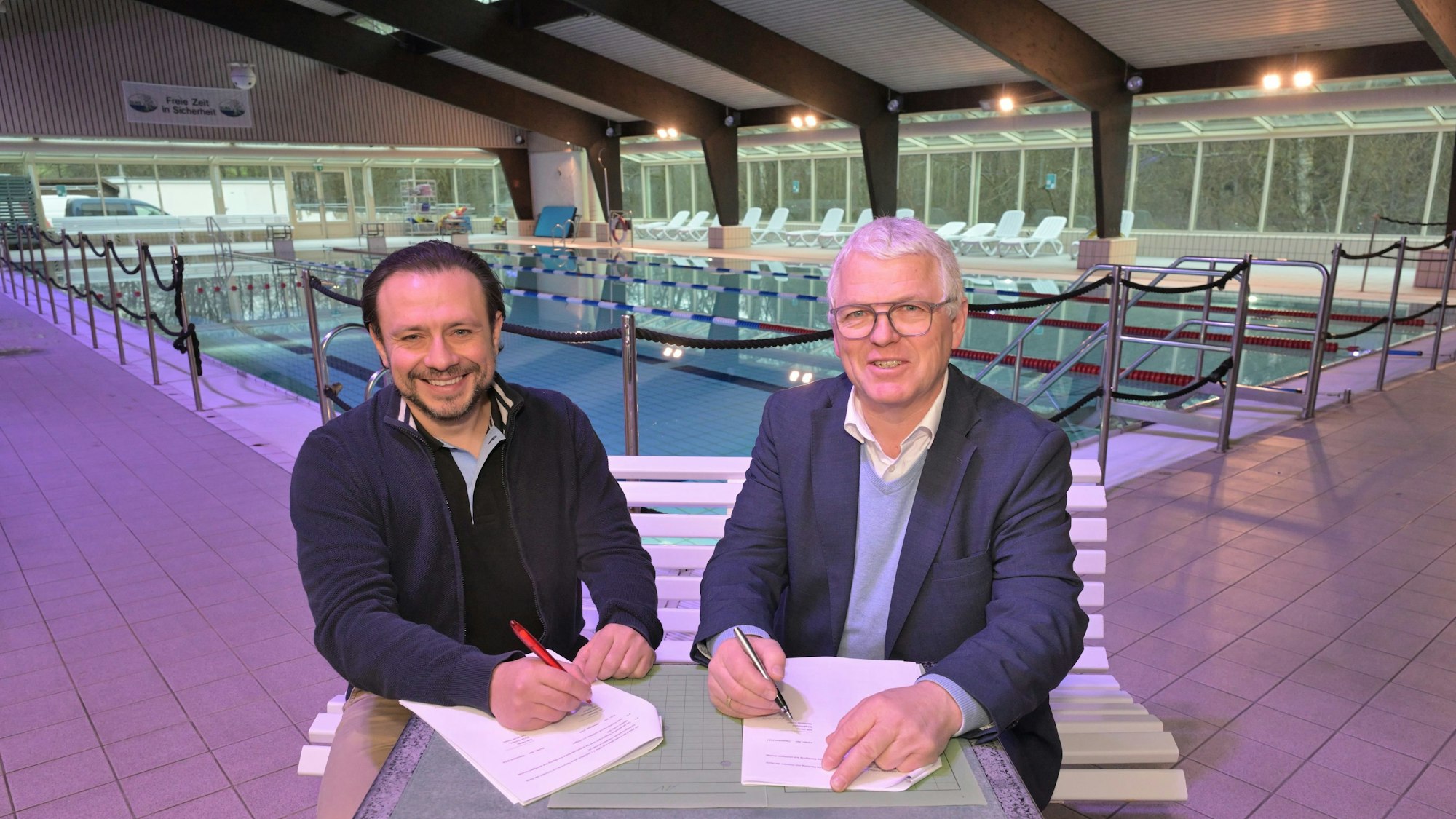 Zwei Männer sitzen in einem Hallenbad an einem Tisch und unterzeichnen einen Vertrag.
