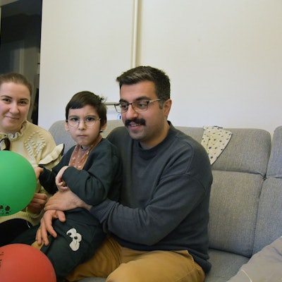 Ein Kind auf dem Schoß seines Vaters, daneben auf dem Sofa die Mutter, im Vordergrund bunte Ballons.