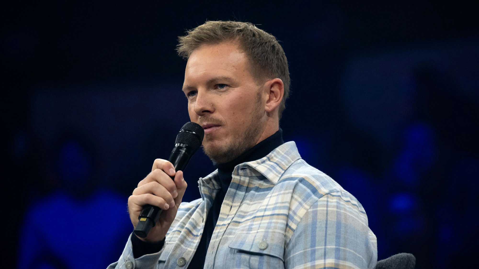 Julian Nagelsmann sitzt mit einem Handmikrofon auf einer Bühne.