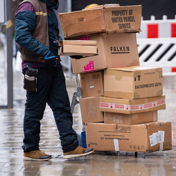 Ein Zusteller steht neben einer Sackkarre, auf der sich Pakete stapeln.