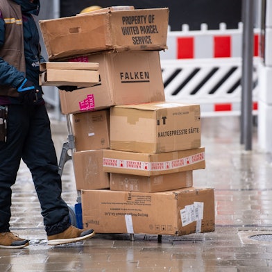 Ein Zusteller steht neben einer Sackkarre, auf der sich Pakete stapeln.