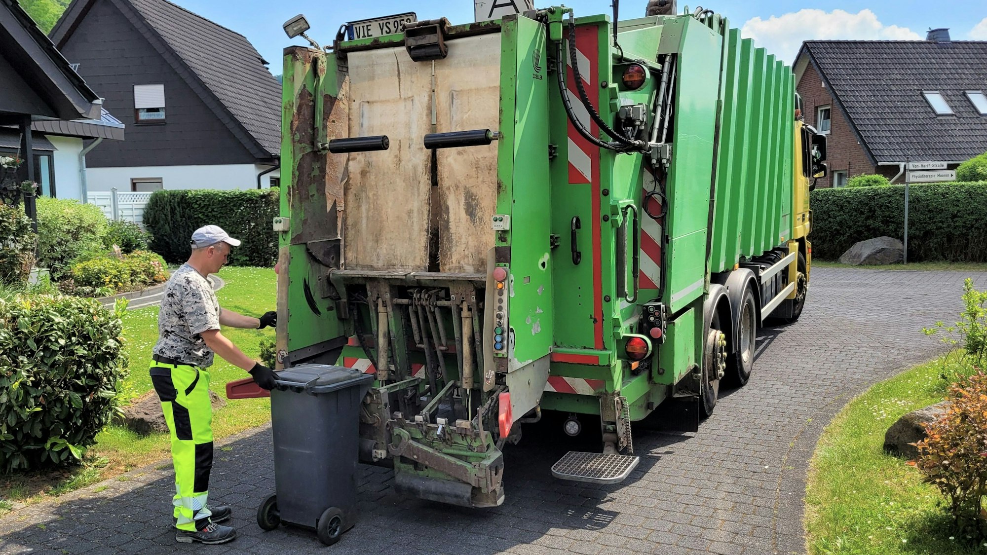 Ein Mitarbeiter führt eine graue Restmülltonne zu einem Müllwagen, der in einem Wohngebiet steht.