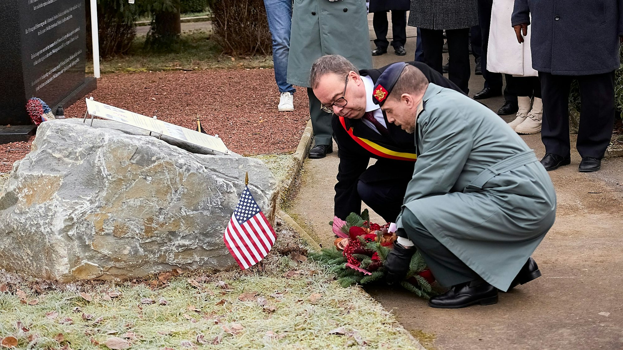 Die beiden Männer knien nieder und legen einen Kranz vor dem Stein mit der Tafel ab.