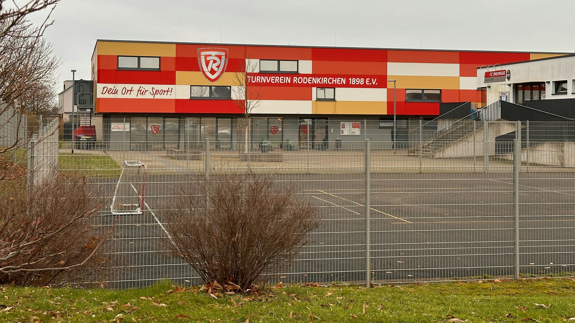 Zu sehen ist das umzäunte rot-orange-gelb-weiße Gebäude des Turnvereins Rodenkirchen.